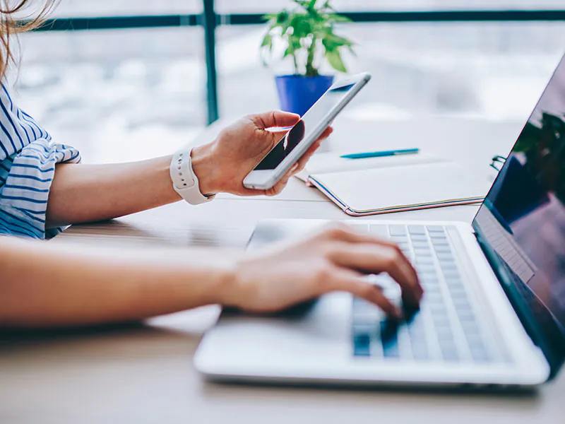 Frau arbeitet an einem Laptop und h&auml;lt ihr Smartphone in der linken Hand.